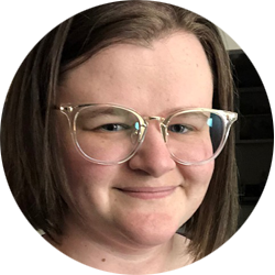 The image features a headshot of a woman with a pleasant expression. She has short, straight brown hair that neatly frames her face. Her complexion is fair, and she is wearing glasses with large, translucent frames that complement her features. She seems to have minimal makeup, giving a natural and approachable look. The woman is smiling subtly at the camera, and the background of the photo is indistinct, keeping the focus on her. The lighting of the photo is soft, casting gentle shadows and giving the image a warm atmosphere. Her attire is not visible in the shot, which focuses on her from the shoulders up.