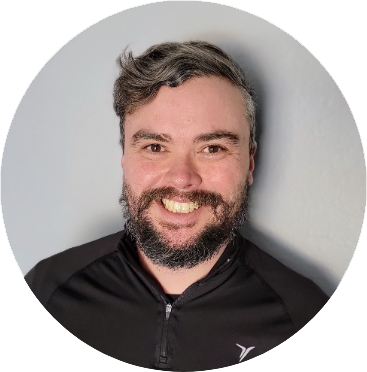 The image is a headshot of a man with a cheerful expression. He has short, wavy, dark hair with a touch of grey, and a well-groomed beard and mustache. His complexion is fair, and he seems to be in a good mood, evidenced by his broad smile showing his teeth. He is wearing a black sports jacket with a zipper, which suggests a casual or sporty attire. The background is a plain light color, which nicely frames the subject. The lighting appears to be from the front, illuminating his face evenly, and enhancing his friendly and relaxed appearance.